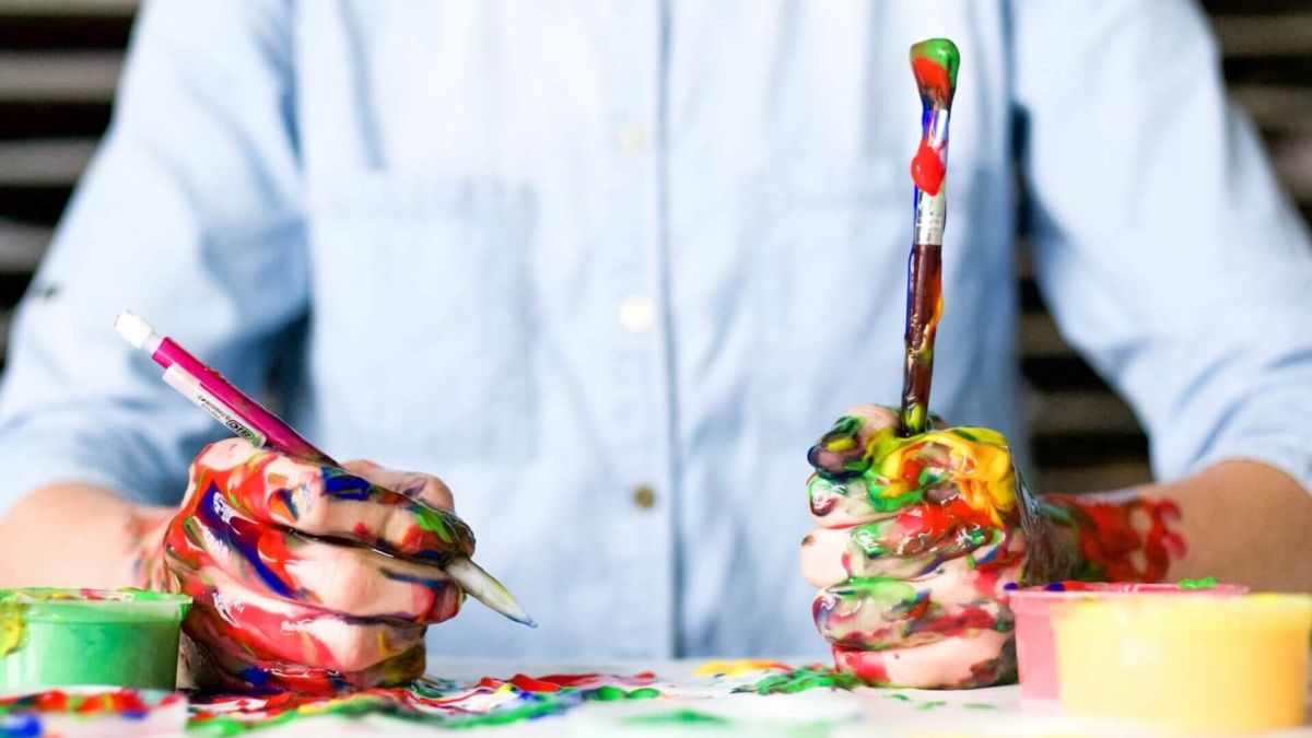 A person is holding a pencil and paint brush, and his hands and its surroundings are painted with mixed color of paint.